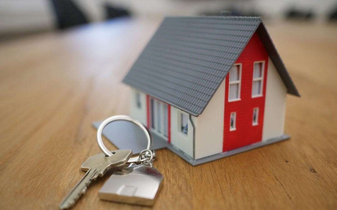 Na drewnianym stole znajdują się miniaturka domu oraz klucze.