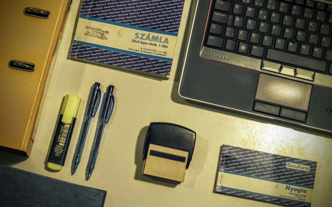 Na stole leżą laptop, zeszyt, druki KP, pieczątka i długopisy oraz zakreślacz.
